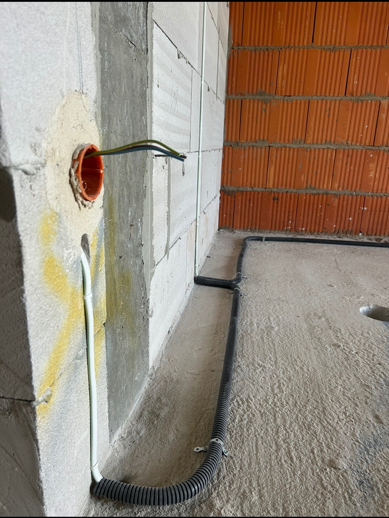 Fragment wnętrza w trakcie prac: przewody w peszlu ułożone wzdłuż ściany z bloczków, puszka instalacyjna z wystającymi kablami. Widoczne mocowania rur do podłoża.