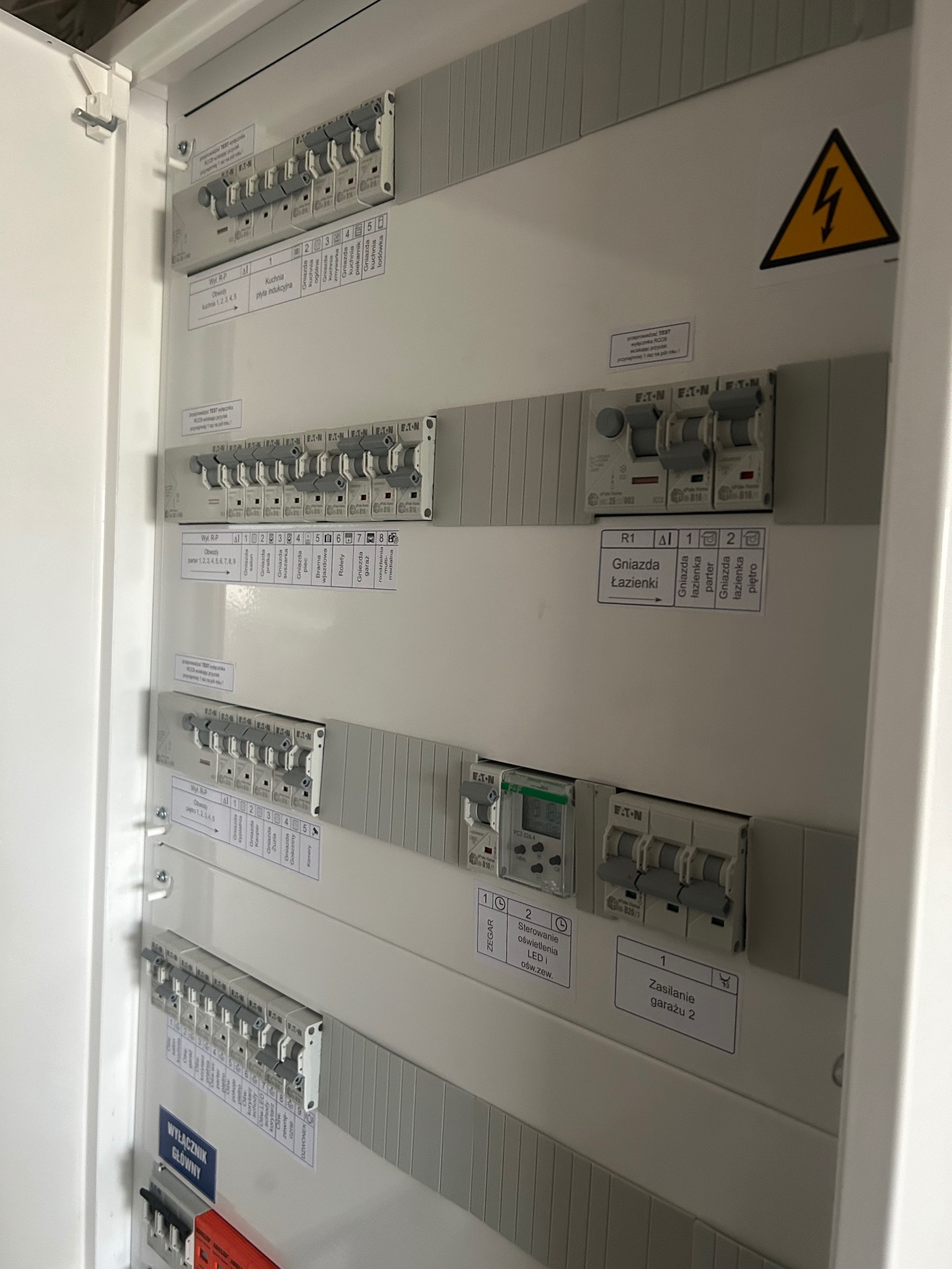 Rozdzielnica elektryczna z bezpiecznikami i opisami obwodów, widoczne zabezpieczenia Eaton, etykiety z nazwami pomieszczeń i funkcji. Żółty znak ostrzegawczy.