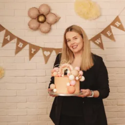 Uśmiechnięta blondynka w czarnym żakiecie trzyma tort z tęczą na tle ceglanej ściany z dekoracjami 'Baby Shower'.