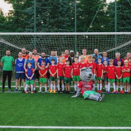 Daria Kriger Photography - Drużyna piłkarska dziecięca z trenerami i maskotką klubową pozuje do zdjęcia na tle bramki i zielonej murawy boiska, w słoneczny dzień.