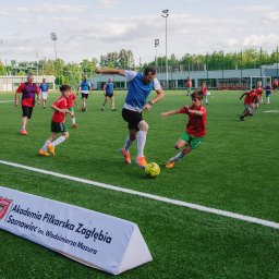 Daria Kriger Photography - Mecz piłki nożnej na zielonym boisku: trener w akcji z młodymi zawodnikami, w tle budynek i drzewa. Na pierwszym planie baner Akademii Piłkarskiej Zagłębia Sosnowiec.