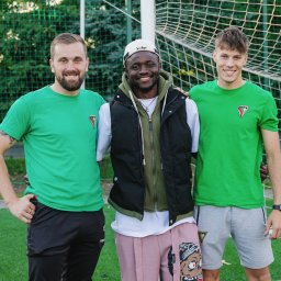 Daria Kriger Photography - Trzech uśmiechniętych mężczyzn w zielonych koszulkach z logo drużyny, stojących na tle siatki bramki na boisku. Jeden w czarnej kamizelce i różowych spodniach.