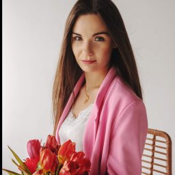 Daria Kriger Photography - Portret kobiety w różowej marynarce, trzymającej bukiet czerwonych tulipanów na jasnym tle. Delikatny uśmiech, złoty naszyjnik i rattanowe krzesło w tle.