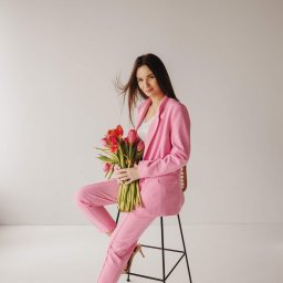 Daria Kriger Photography - Portret kobiety w różowym garniturze, siedzącej na krześle z bukietem czerwonych tulipanów. Jasne tło, naturalne światło, delikatny uśmiech. Elegancka sesja.