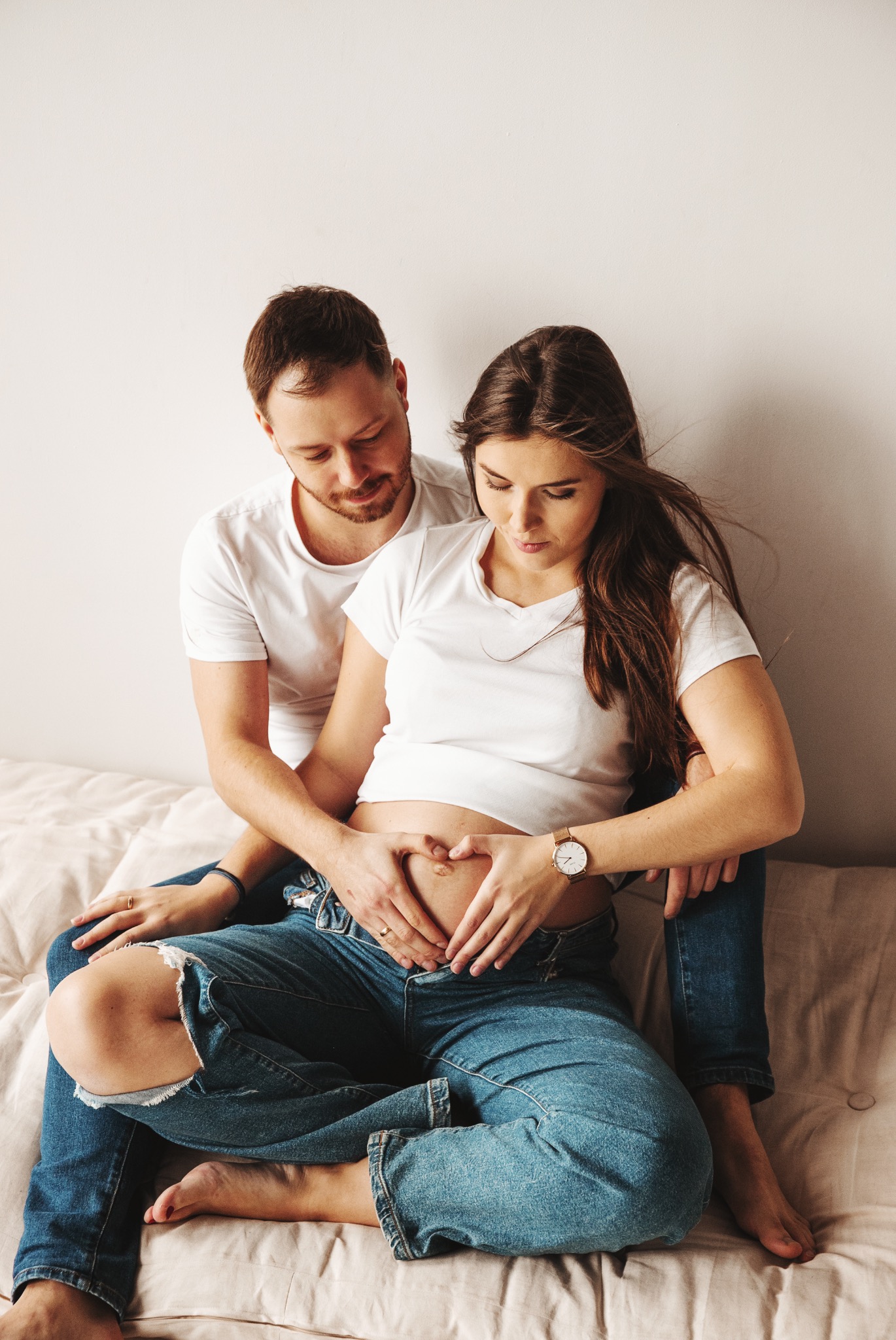 Uśmiechnięta para w jasnych ubraniach siedzi na łóżku. Kobieta w ciąży, z rękami ułożonymi w kształt serca na brzuchu. Naturalne światło.