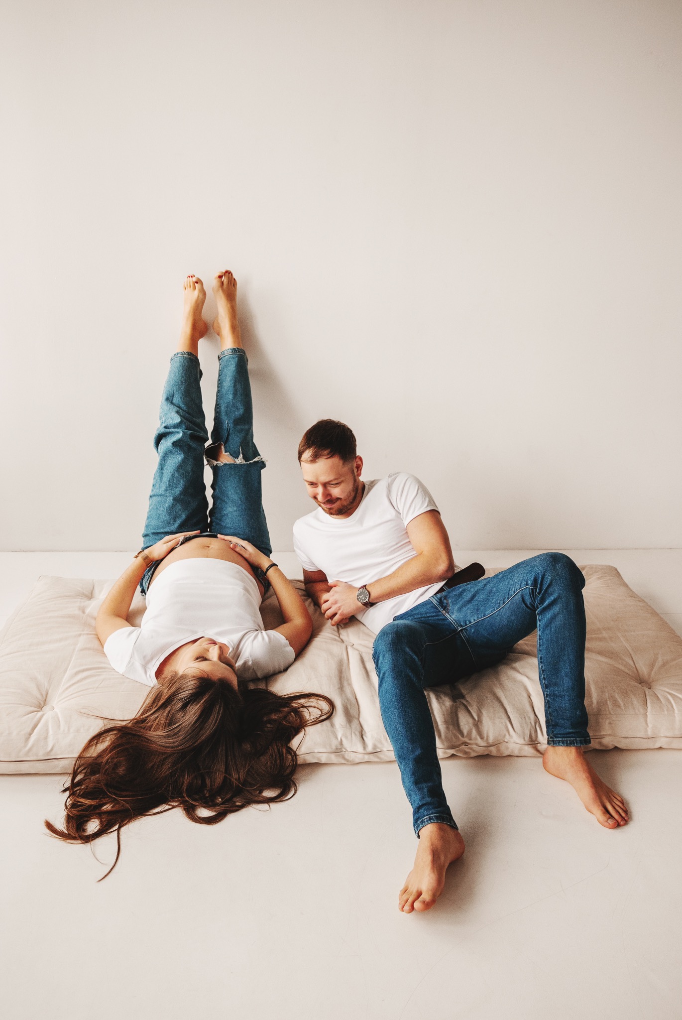 Uśmiechnięty mężczyzna siedzi obok leżącej na plecach kobiety w ciąży, która opiera nogi o białą ścianę. Jasne, minimalistyczne tło, naturalne światło.