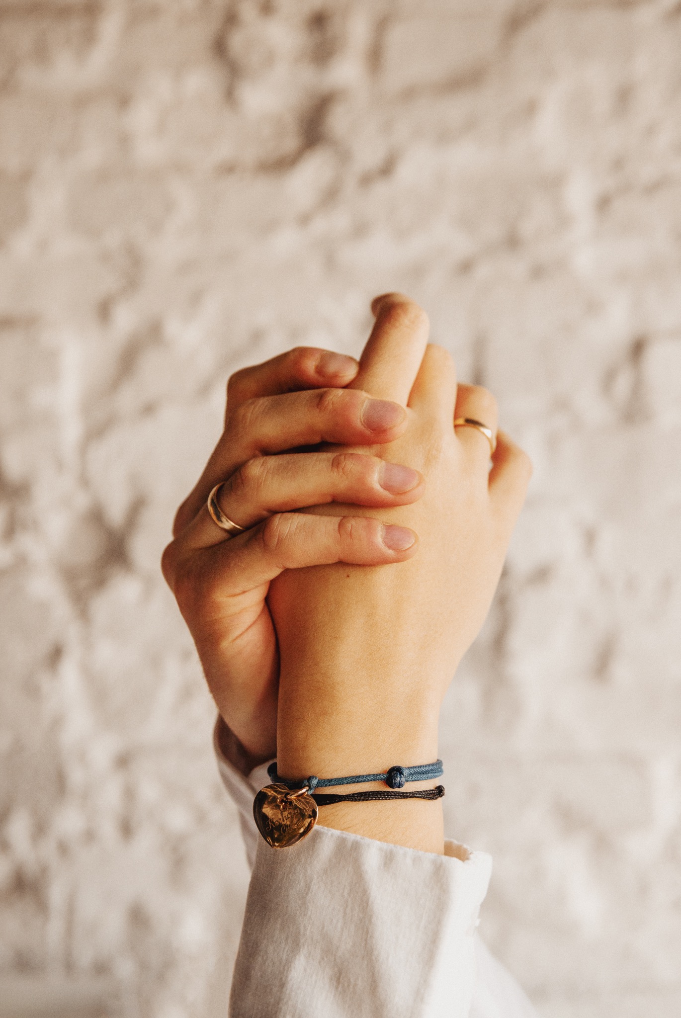 Złączone dłonie z pierścionkami i bransoletkami na tle białej ściany z cegieł. Dłoń z bransoletką z zawieszką w kształcie serca.