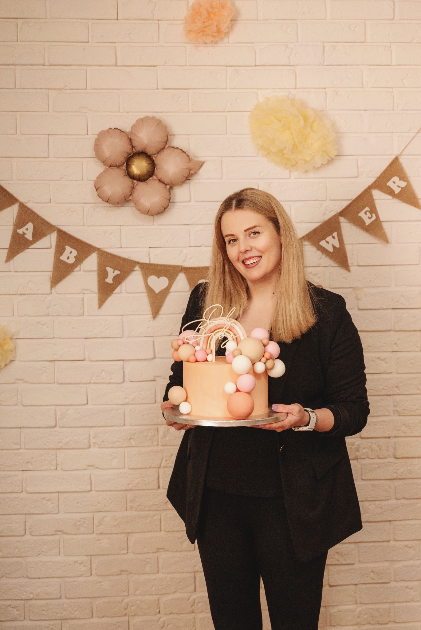 Uśmiechnięta blondynka w czarnym żakiecie trzyma tort z tęczą na tle ceglanej ściany z dekoracjami 'Baby Shower'.