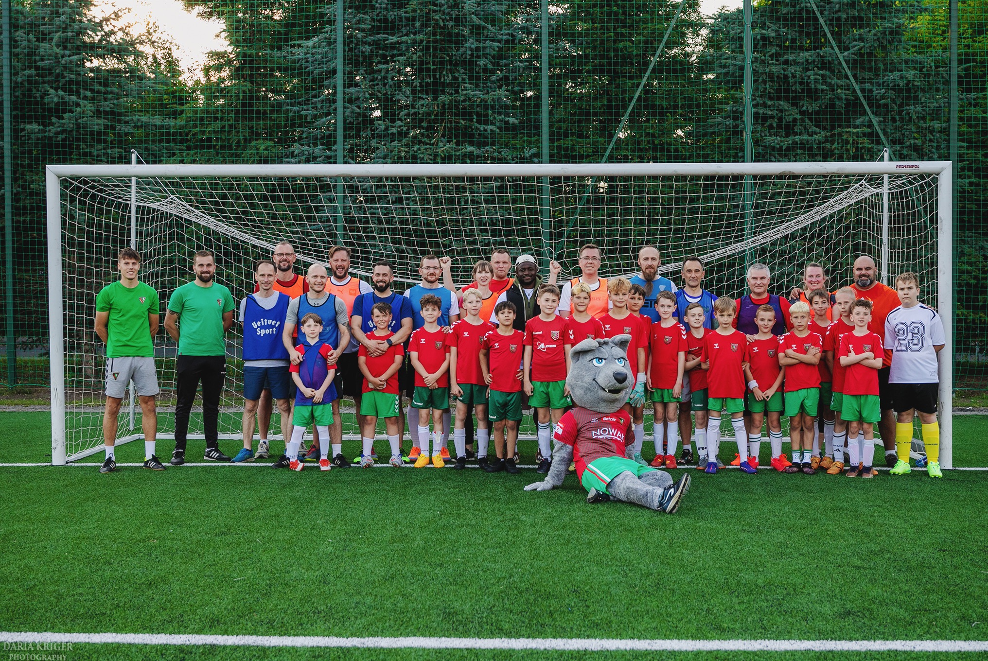 Drużyna piłkarska dziecięca z trenerami i maskotką klubową pozuje do zdjęcia na tle bramki i zielonej murawy boiska, w słoneczny dzień.