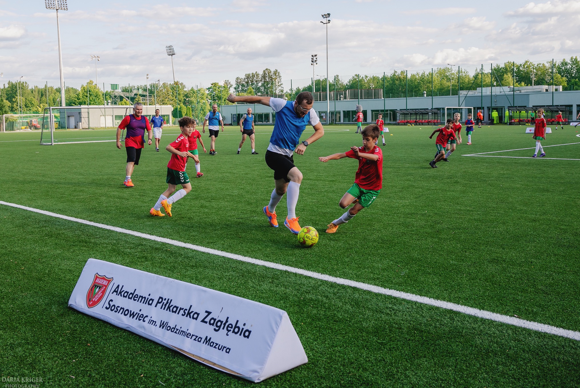 Mecz piłki nożnej na zielonym boisku: trener w akcji z młodymi zawodnikami, w tle budynek i drzewa. Na pierwszym planie baner Akademii Piłkarskiej Zagłębia Sosnowiec.