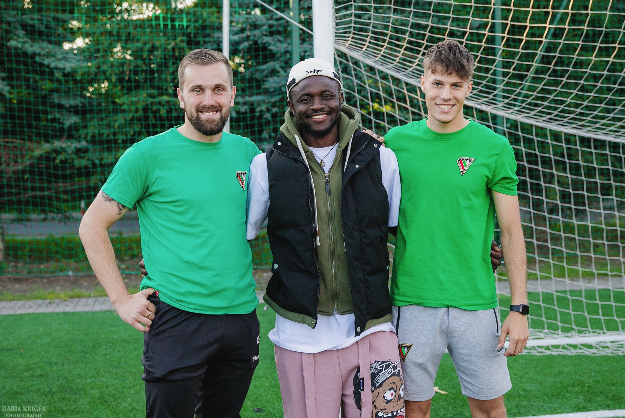 Trzech uśmiechniętych mężczyzn w zielonych koszulkach z logo drużyny, stojących na tle siatki bramki na boisku. Jeden w czarnej kamizelce i różowych spodniach.