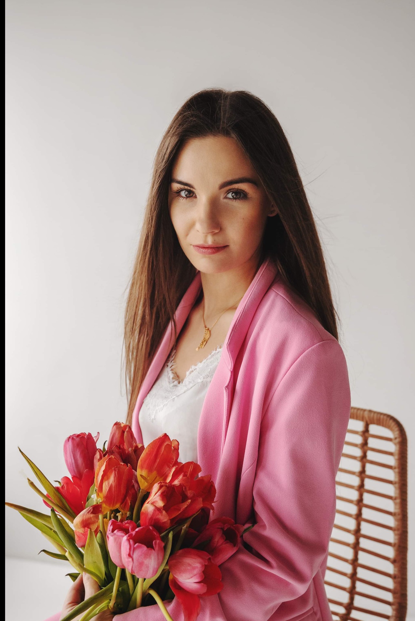 Portret kobiety w różowej marynarce, trzymającej bukiet czerwonych tulipanów na jasnym tle. Delikatny uśmiech, złoty naszyjnik i rattanowe krzesło w tle.