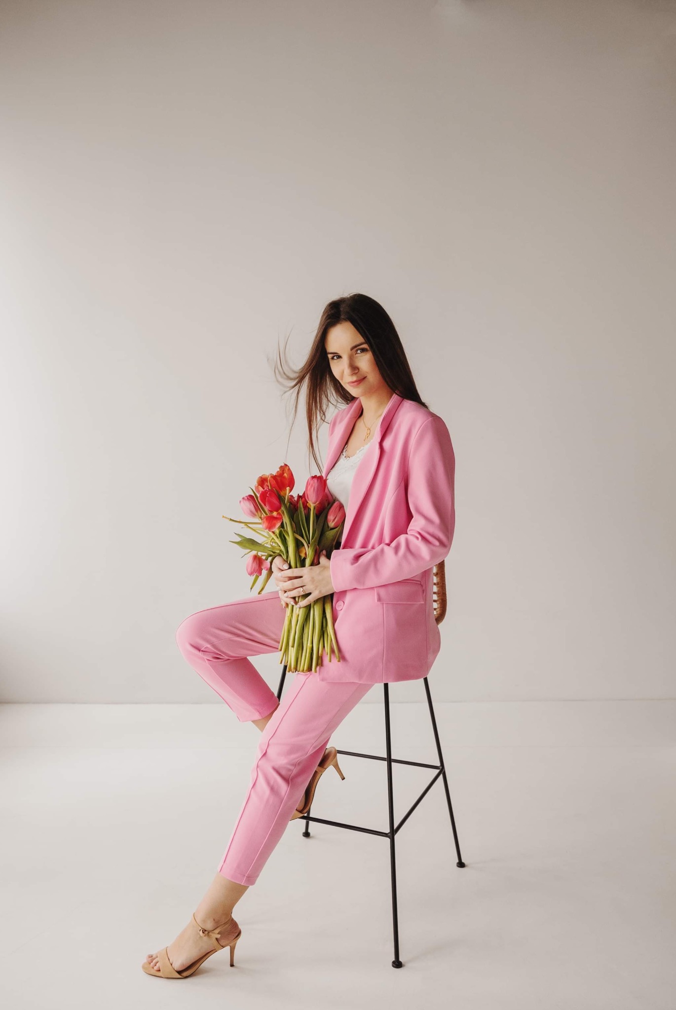 Portret kobiety w różowym garniturze, siedzącej na krześle z bukietem czerwonych tulipanów. Jasne tło, naturalne światło, delikatny uśmiech. Elegancka sesja.