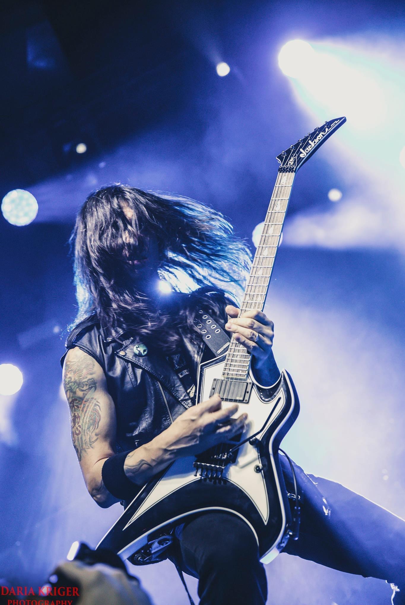 Dynamiczne ujęcie gitarzysty w skórzanej kamizelce, z tatuażem na ramieniu, podczas koncertu w niebieskim oświetleniu, z rozwianymi włosami i gitarą Jackson.