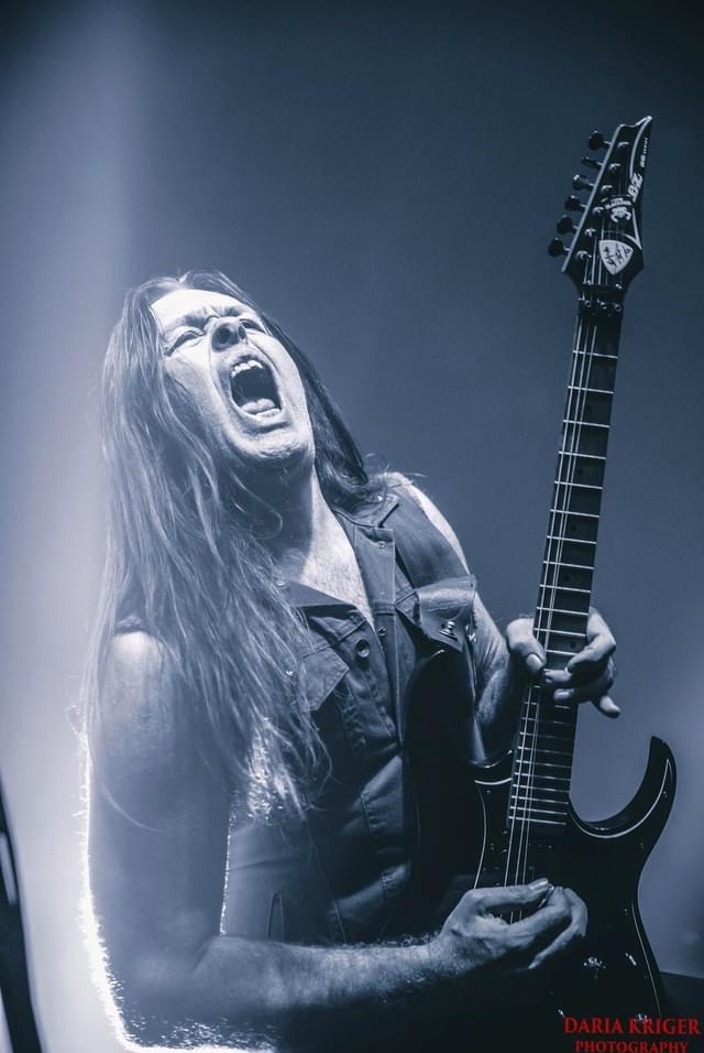 Ekspresyjne zdjęcie muzyka z długimi włosami, grającego na czarnej gitarze elektrycznej podczas koncertu, z otwartymi ustami i intensywnym oświetleniem.