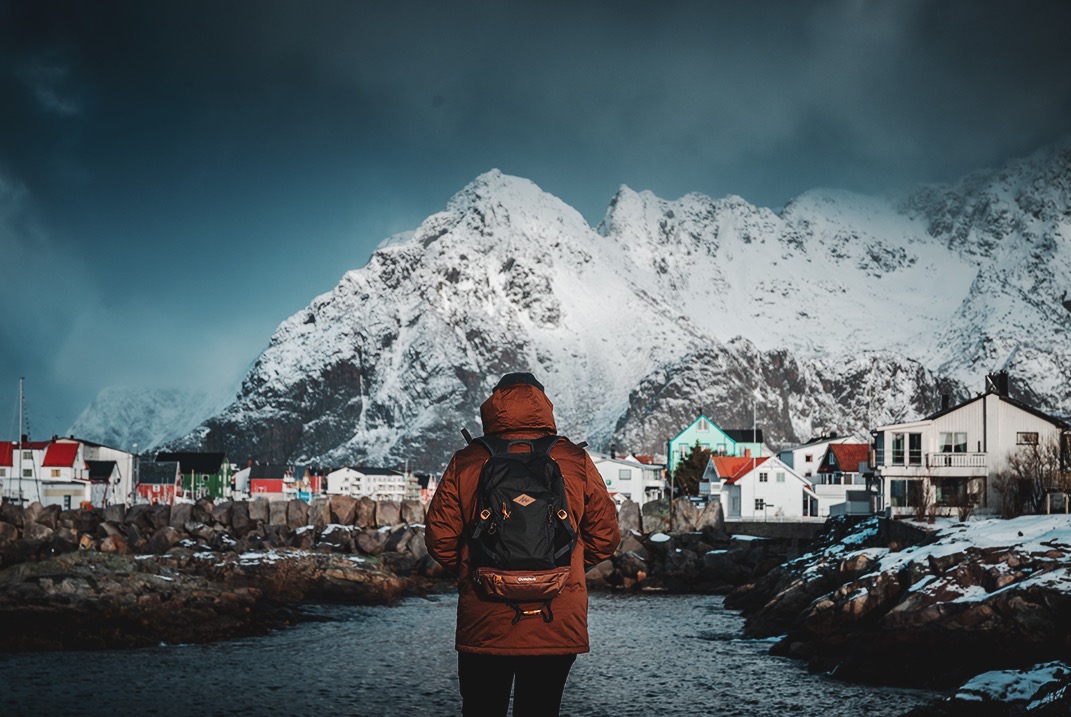 Osoba z plecakiem podziwia zimowy krajobraz w Sarnowie: ośnieżone góry i kolorowe domki rybackie pod pochmurnym niebem.