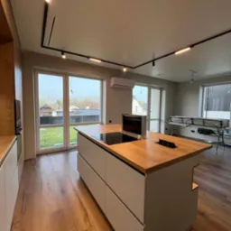 Nowoczesna wyspa kuchenna z drewnianym blatem i indukcyjną płytą, minimalistyczne szare fronty, oświetlenie szynowe, widok na ogród przez duże okna.