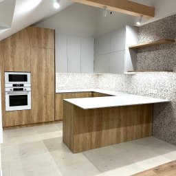 TTM - Nowoczesna kuchnia z drewnianymi frontami i białymi szafkami, wyspa kuchenna z białym blatem, oświetlenie punktowe na drewnianej belce stropowej.