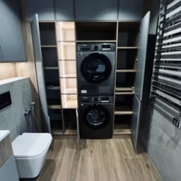 Szara zabudowa meblowa w łazience z pralką i suszarką Samsung, otwarte p&oacute;łki z oświetleniem LED, płytki imitujące drewno na podłodze, grzejnik drabinkowy.