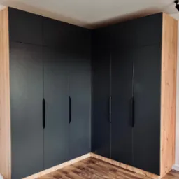 Duża, narożna szafa wnękowa z czarnymi frontami i drewnianym korpusem, dopasowana do podłogi. Minimalistyczne uchwyty, oświetlenie sufitowe.