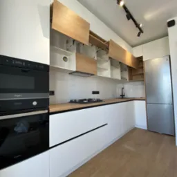 Nowoczesna, jasna kuchnia z białymi frontami i drewnianymi akcentami. Górne szafki otwarte, oświetlenie szynowe. Minimalistyczny design z czarnymi detalami.