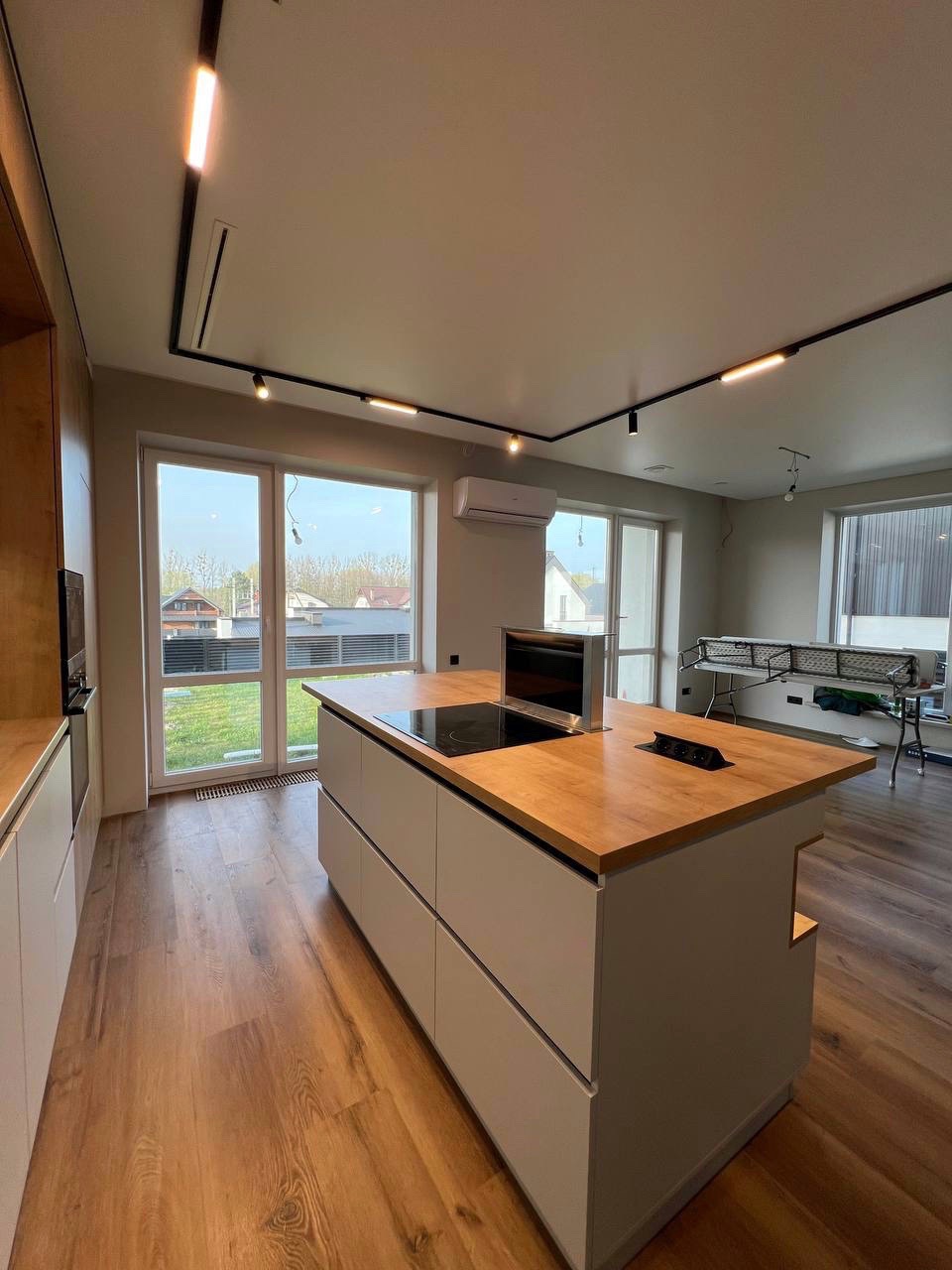 Nowoczesna wyspa kuchenna z drewnianym blatem i indukcyjną płytą, minimalistyczne szare fronty, oświetlenie szynowe, widok na ogród przez duże okna.
