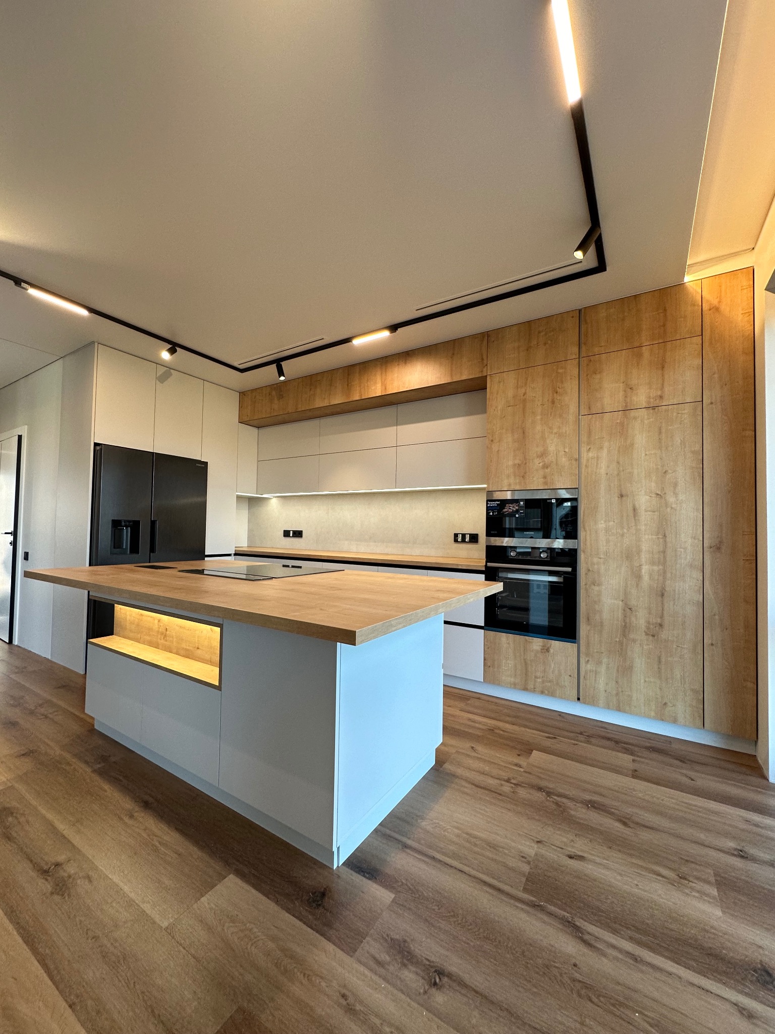 Nowoczesna kuchnia we Wrocławiu: wyspa z drewnianym blatem, jasne szafki i oświetlenie punktowe. Zabudowa z piekarnikiem i lodówka w czarnym kolorze.