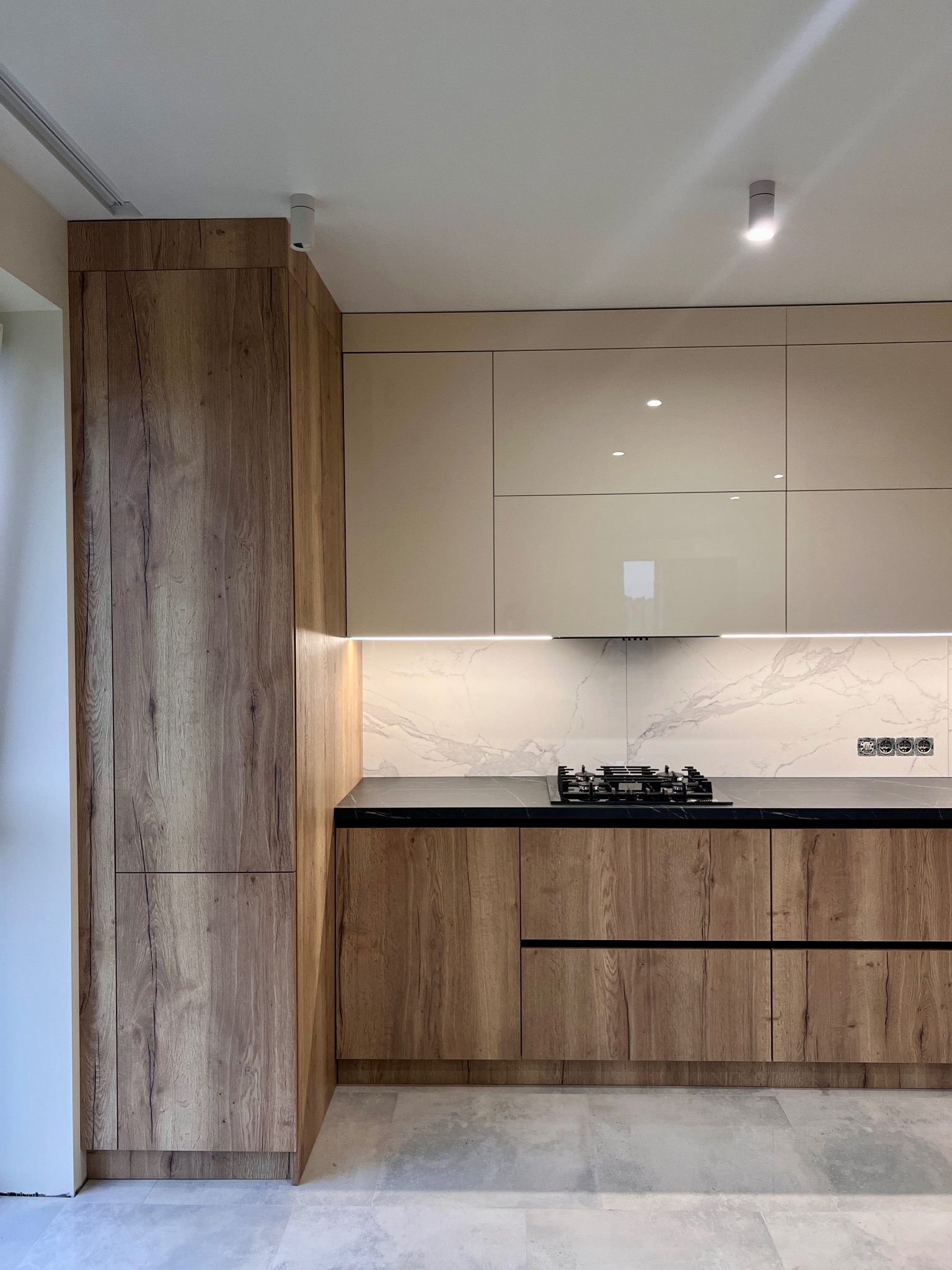 Nowoczesna kuchnia: drewniane szafki dolne z czarnym blatem, beżowe szafki górne z białym marmurowym panelem. Oświetlenie LED podkreśla elegancję.