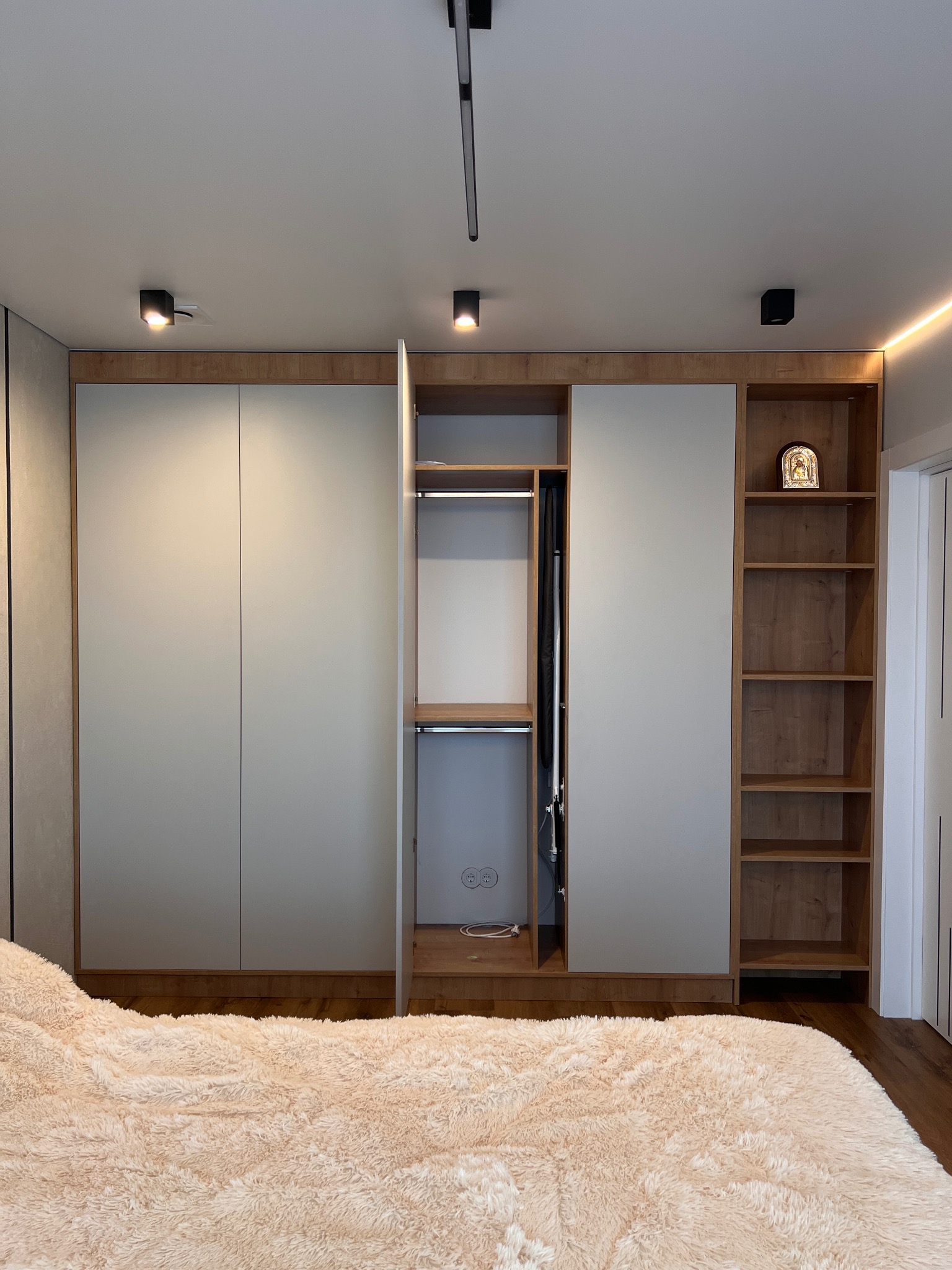 Nowoczesna szafa wnękowa z drewnianymi elementami i szarymi frontami, częściowo otwarta, z oświetleniem punktowym na suficie i dekoracyjną ikoną na półce.