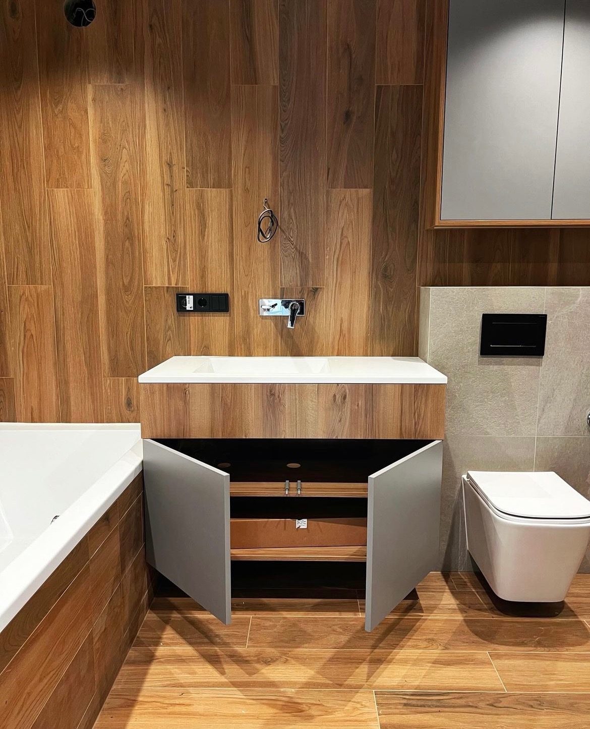 Nowoczesna łazienka z drewnianą zabudową umywalki z otwartymi drzwiczkami, płytki imitujące drewno na ścianie i podłodze, biała wanna i toaleta.