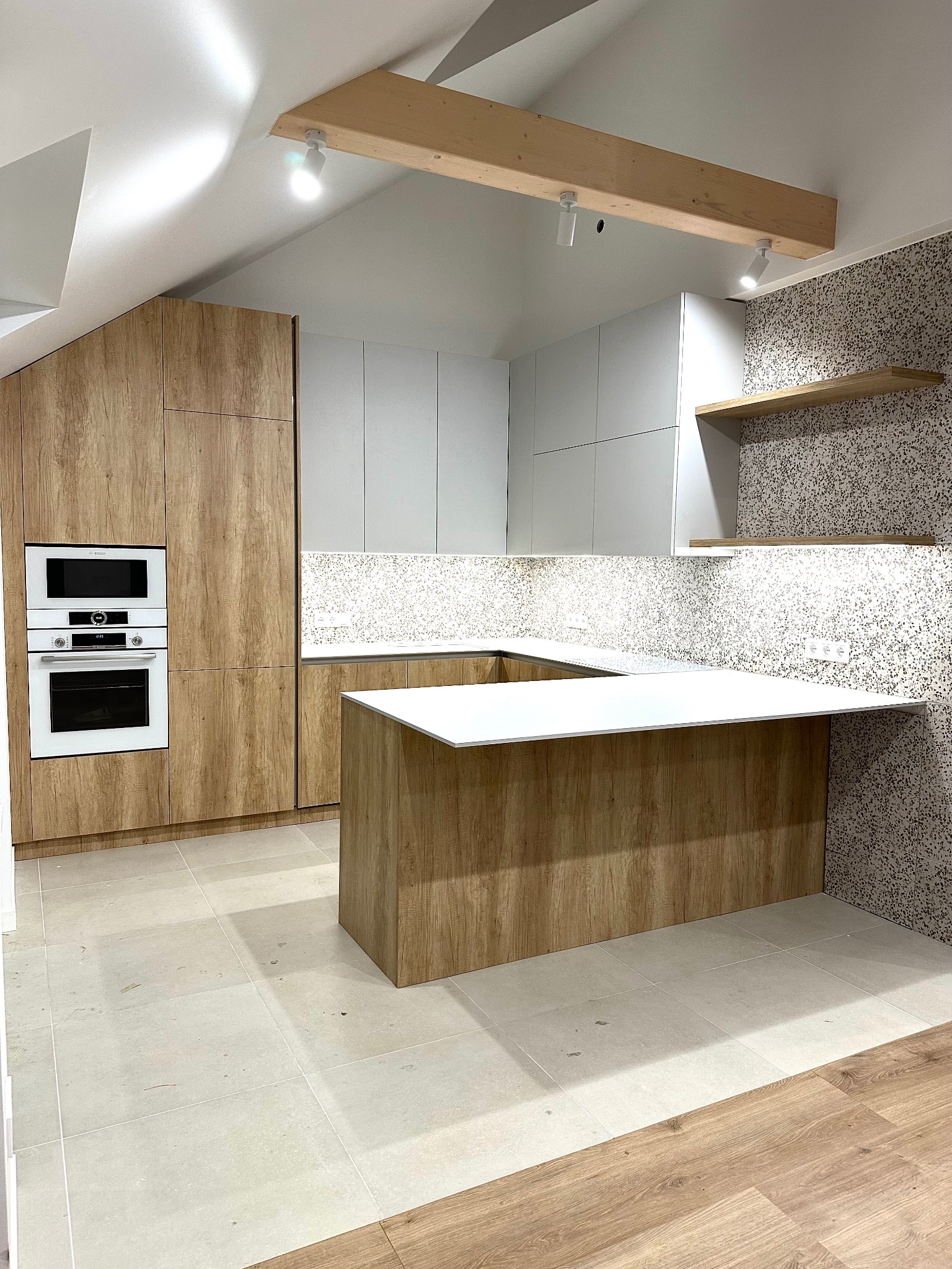 Nowoczesna kuchnia z drewnianymi frontami i białymi szafkami, wyspa kuchenna z białym blatem, oświetlenie punktowe na drewnianej belce stropowej.