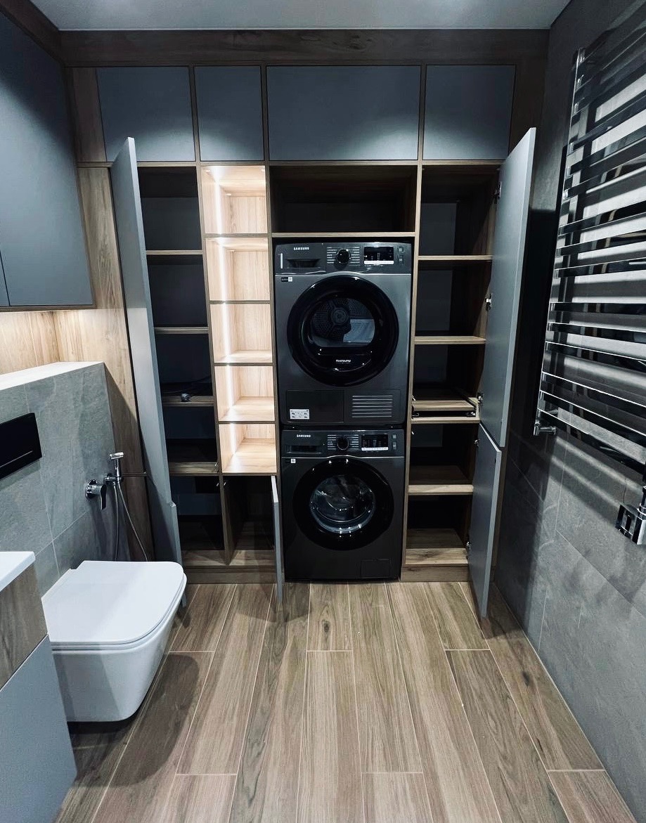 Szara zabudowa meblowa w łazience z pralką i suszarką Samsung, otwarte półki z oświetleniem LED, płytki imitujące drewno na podłodze, grzejnik drabinkowy.
