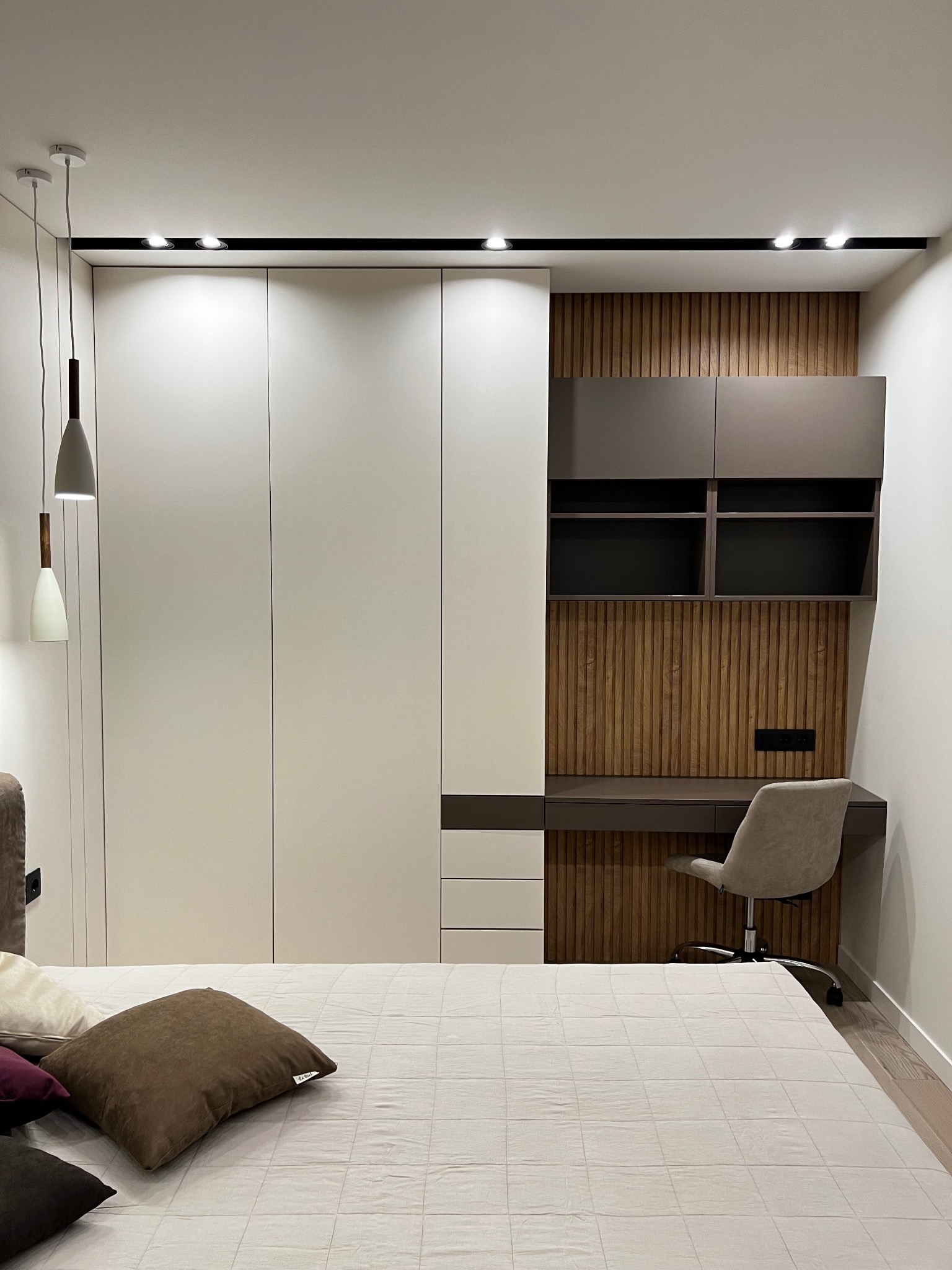 Nowoczesna szafa wnękowa z biurkiem i półkami w sypialni. Minimalistyczny design, białe fronty, drewniane akcenty, oświetlenie punktowe i wiszące lampy.
