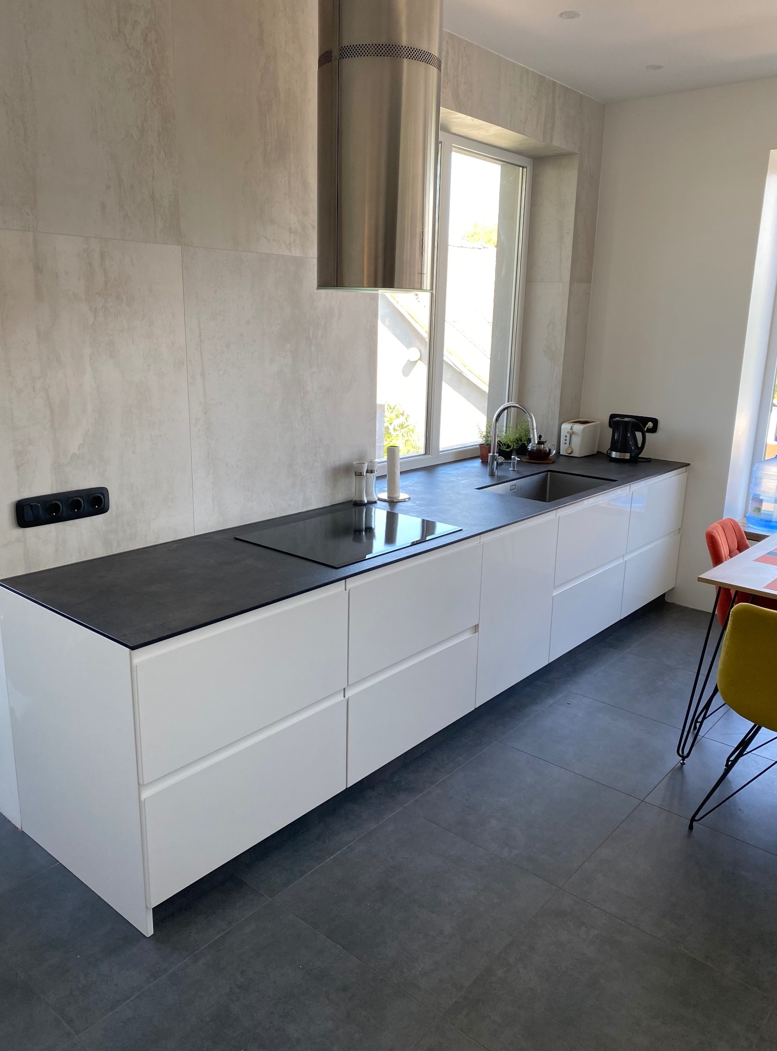 Nowoczesna kuchnia z białymi szafkami i czarnym blatem. Nad blatem stalowy okap i okno z widokiem na zewnątrz. Minimalistyczny design, szare płytki na podłodze.