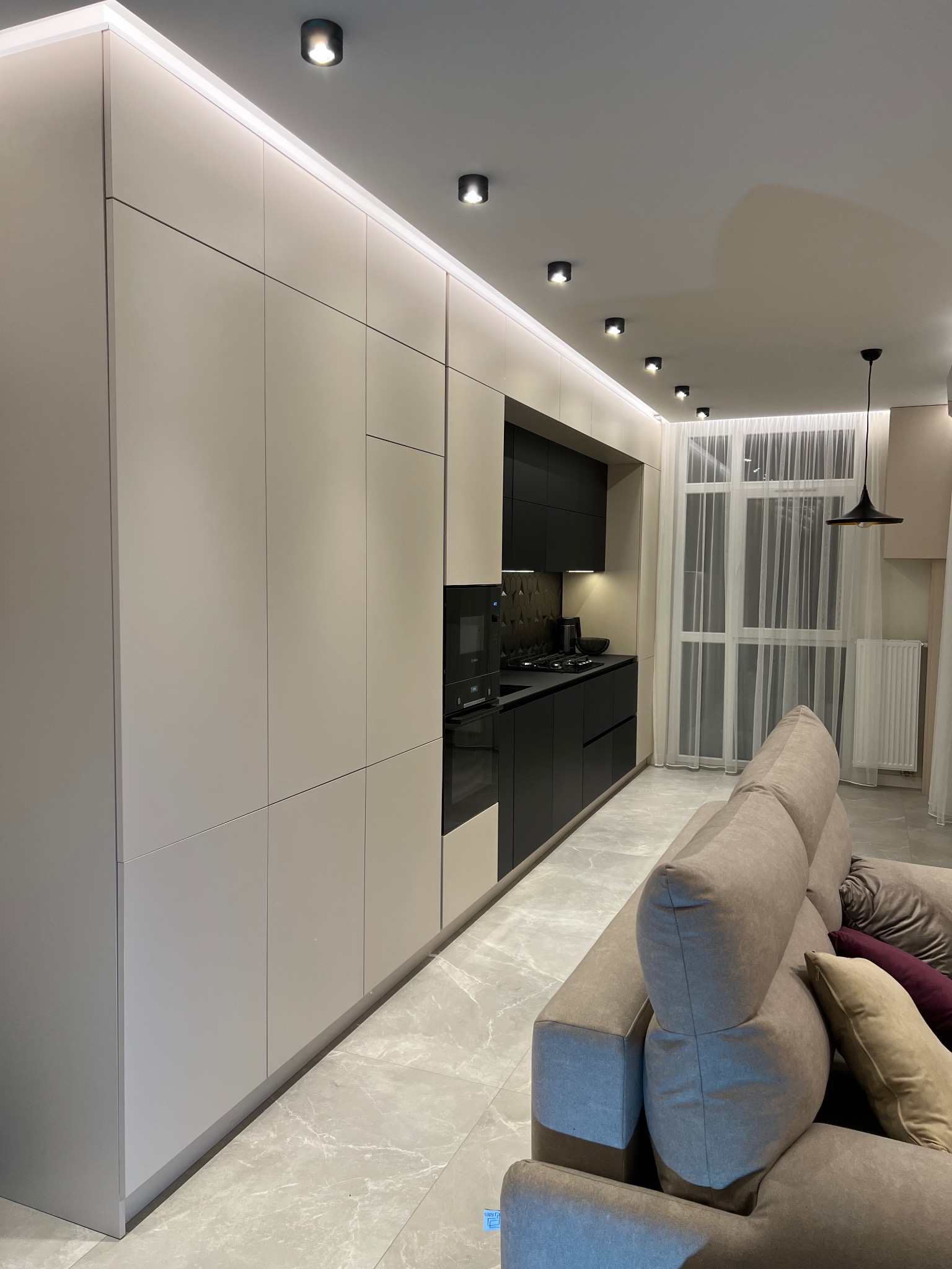 Nowoczesna kuchnia z beżowymi szafkami, czarnymi akcentami i oświetleniem LED. Widok z salonu z beżową sofą i marmurową podłogą.