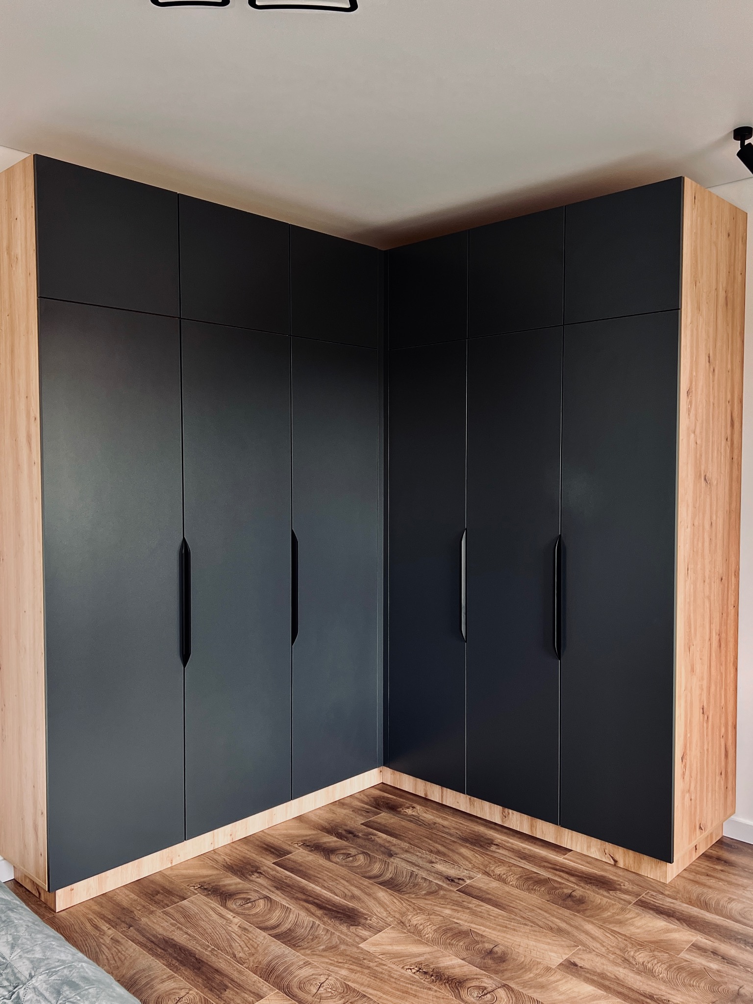 Duża, narożna szafa wnękowa z czarnymi frontami i drewnianym korpusem, dopasowana do podłogi. Minimalistyczne uchwyty, oświetlenie sufitowe.