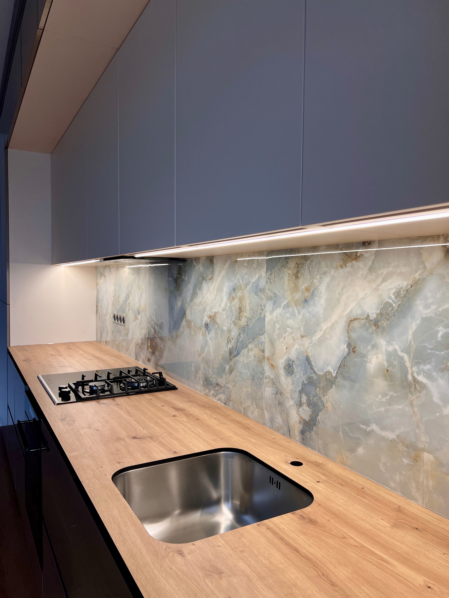 Nowoczesna kuchnia: drewniany blat z wbudowanym zlewem i płytą gazową, marmurowy panel ścienny podświetlony LED, szare szafki wiszące.