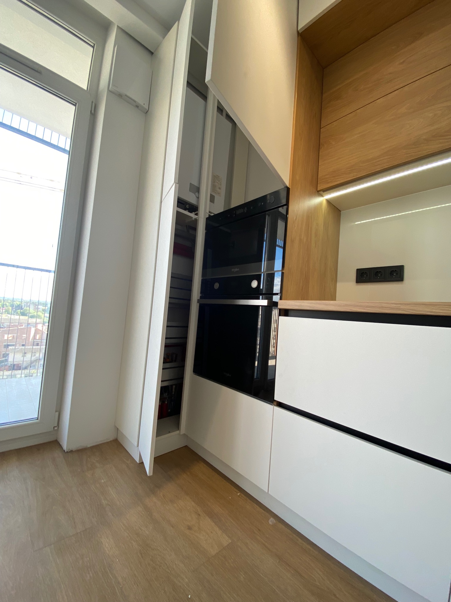 Nowoczesna kuchnia: zabudowa z piekarnikiem i szufladami cargo, białe fronty, drewniane akcenty, oświetlenie LED, widok na okno i fragment zabudowy przy oknie.