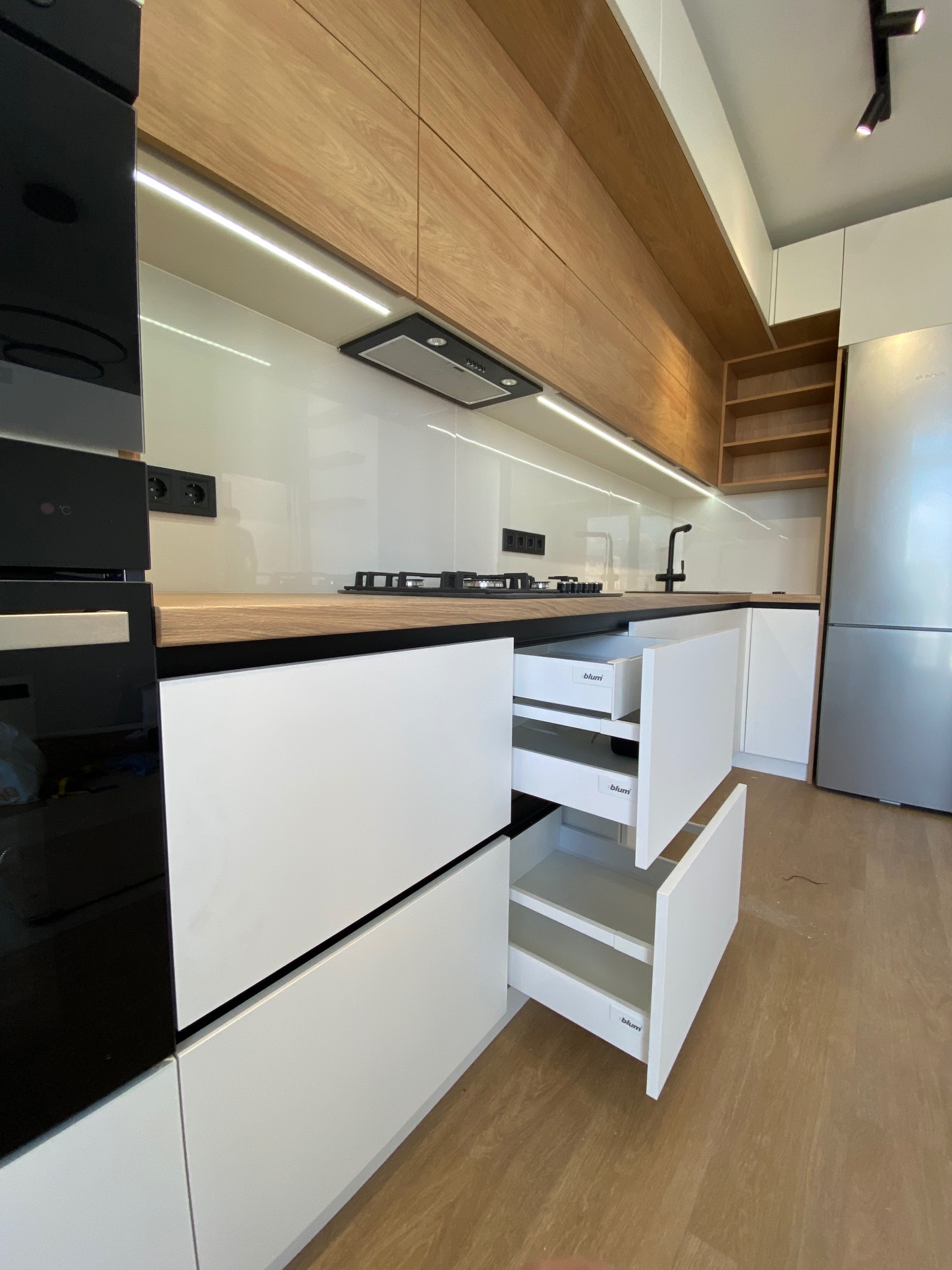 Nowoczesna kuchnia z białymi szafkami i drewnianymi górnymi szafkami. Dwie szuflady są otwarte, ukazując system Blum. Widoczny blat drewniany i płyta gazowa.