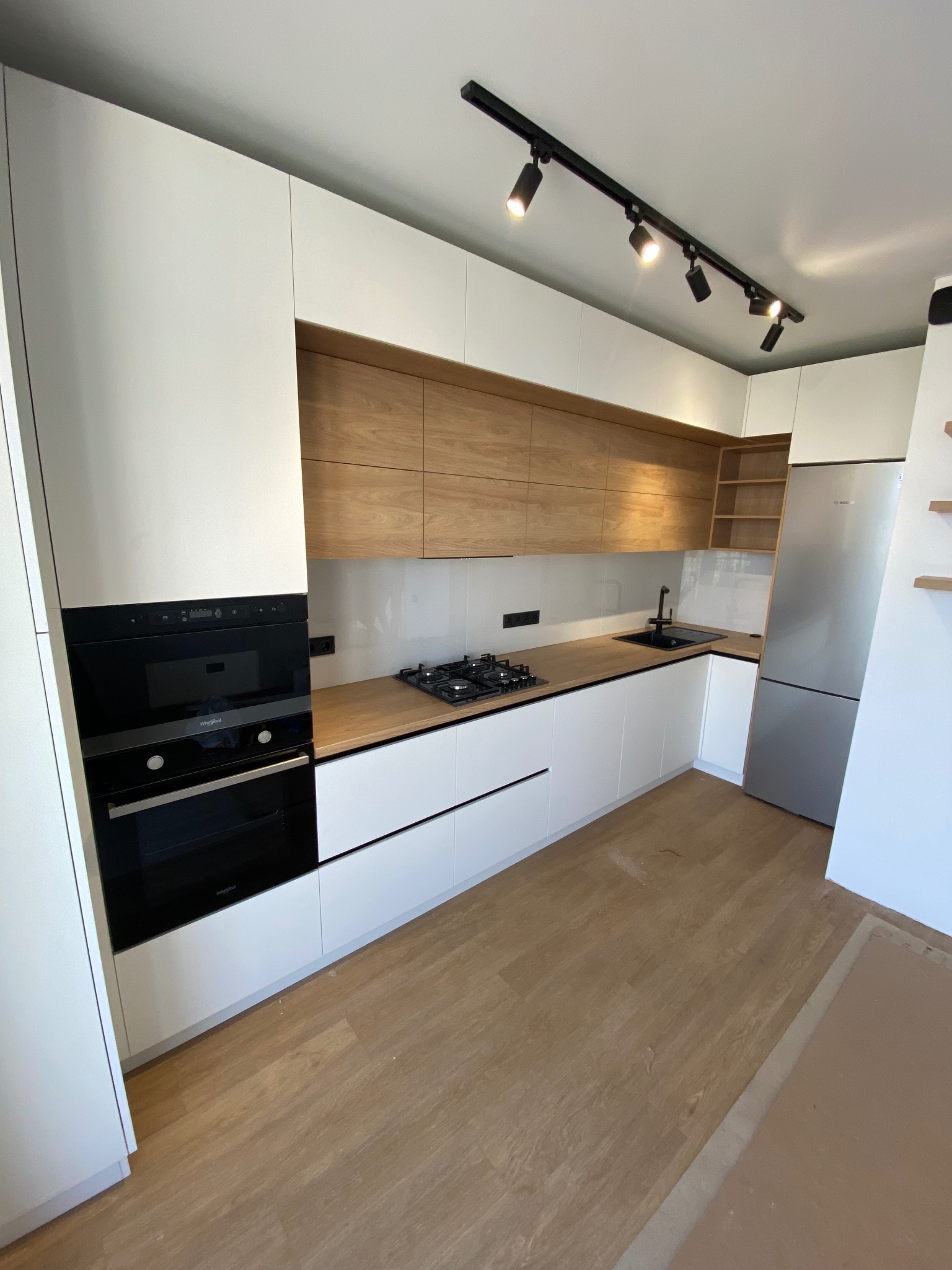 Nowoczesna kuchnia z białymi szafkami, drewnianymi akcentami i czarnymi detalami, oświetlona reflektorami sufitowymi. Widoczny sprzęt AGD i drewniana podłoga.