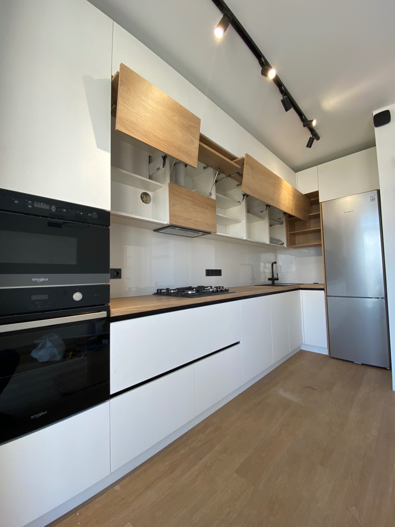 Nowoczesna, jasna kuchnia z białymi frontami i drewnianymi akcentami. Górne szafki otwarte, oświetlenie szynowe. Minimalistyczny design z czarnymi detalami.