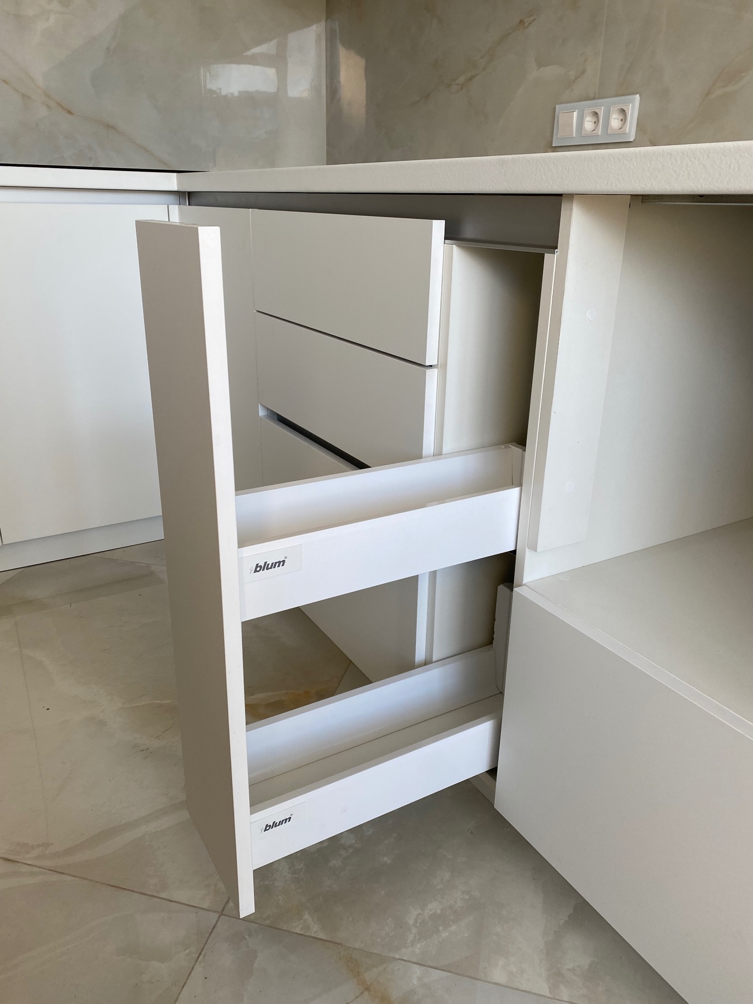 Biała szafka kuchenna z wysuwanym cargo marki Blum, widoczne półki i front. Minimalistyczny design, jasne wnętrze, marmurowe płytki w tle. Gniazdko elektryczne nad blatem.