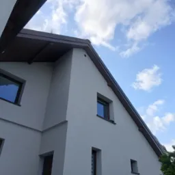 Jasny, dwukondygnacyjny dom z szarym tynkiem, brązowym dachem i oknami. Widok z dołu na elewację i fragment nieba z chmurami. Nowoczesna architektura.