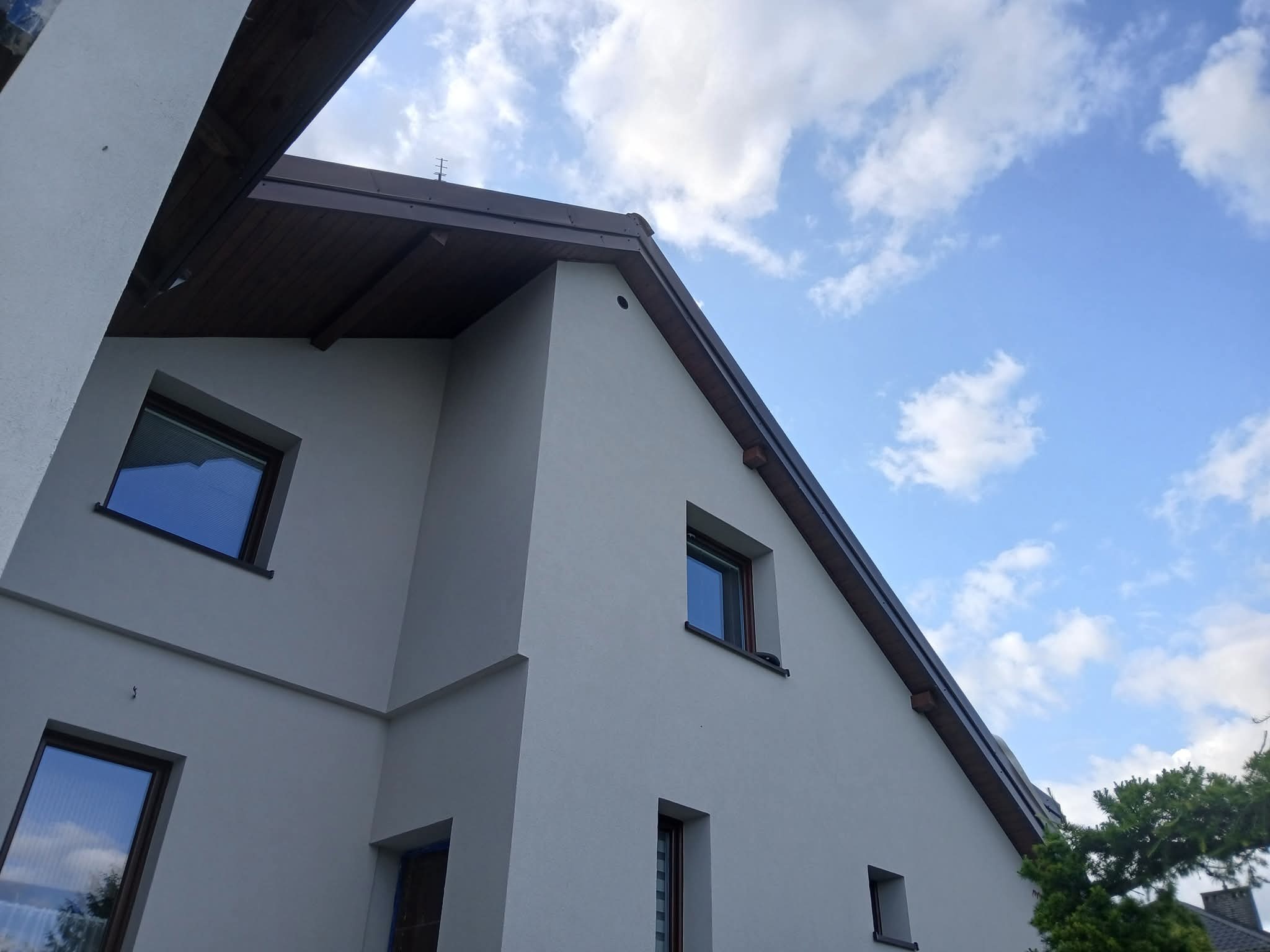 Jasny, dwukondygnacyjny dom z szarym tynkiem, brązowym dachem i oknami. Widok z dołu na elewację i fragment nieba z chmurami. Nowoczesna architektura.