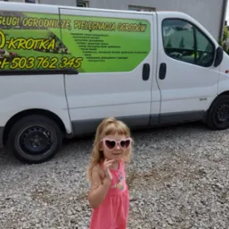 Biały bus z reklamą usług ogrodniczych '100-Krotka' i numerem telefonu, na tle żwirowej powierzchni i drewnianej altany. Na pierwszym planie dziewczynka w różowych okularach.