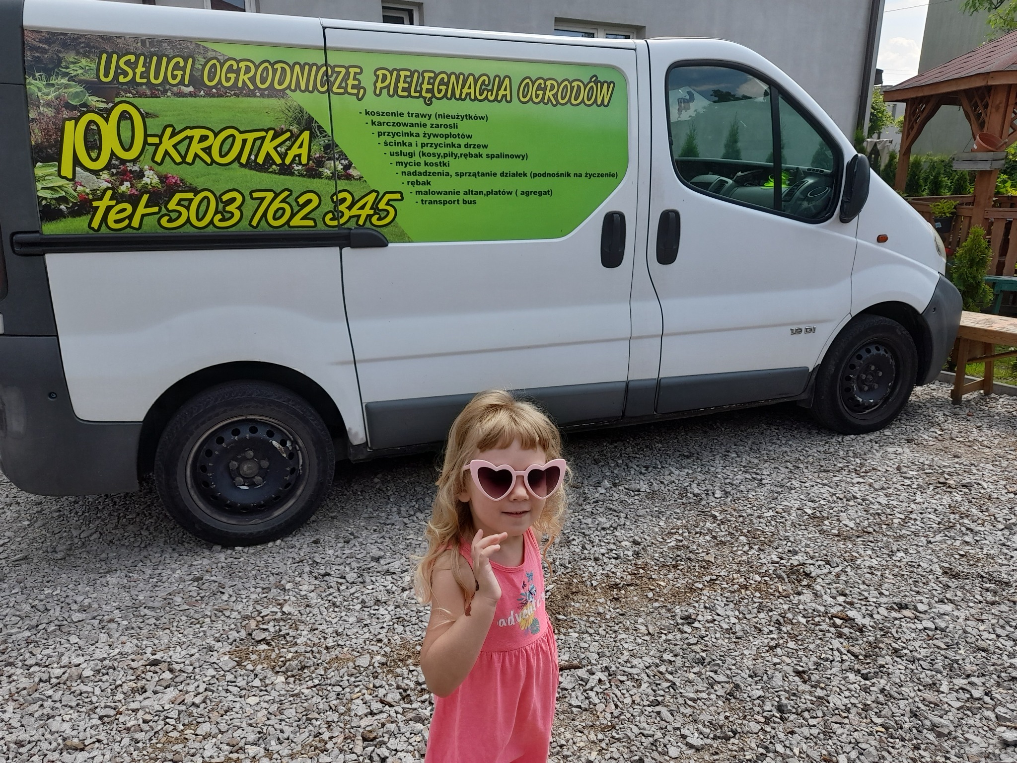 Biały bus z reklamą usług ogrodniczych '100-Krotka' i numerem telefonu, na tle żwirowej powierzchni i drewnianej altany. Na pierwszym planie dziewczynka w różowych okularach.