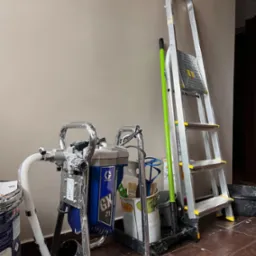 Sprzęt malarski: agregat hydrodynamiczny Graco GX 21, aluminiowa drabina, pędzle i farby, gotowe do użycia przy ścianie w odcieniach beżu.