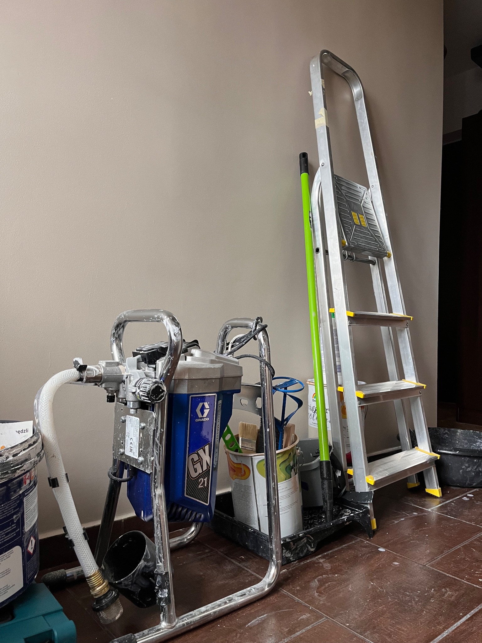 Sprzęt malarski: agregat hydrodynamiczny Graco GX 21, aluminiowa drabina, pędzle i farby, gotowe do użycia przy ścianie w odcieniach beżu.