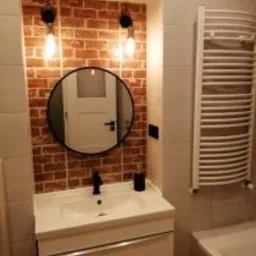 Wykończona łazienka: umywalka z szafką, okrągłe lustro w czarnej ramie, ściana z cegieł, oświetlenie w stylu loft i biały grzejnik drabinkowy.