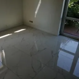 Błyszcząca, marmurowa podłoga z dużych płytek w jasnym pokoju z oknem balkonowym. Widoczne refleksy światła podkreślają wzór i połysk.